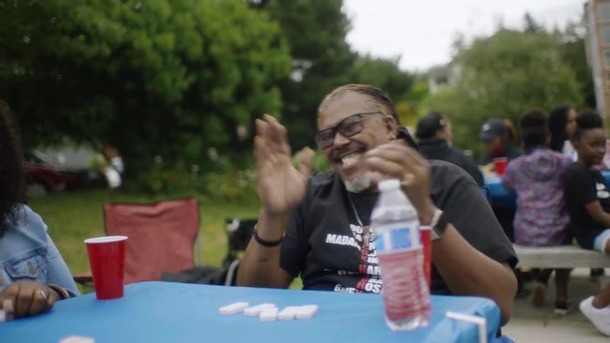 People are sitting at a table outside in a park playing a game of dominoes and laughing