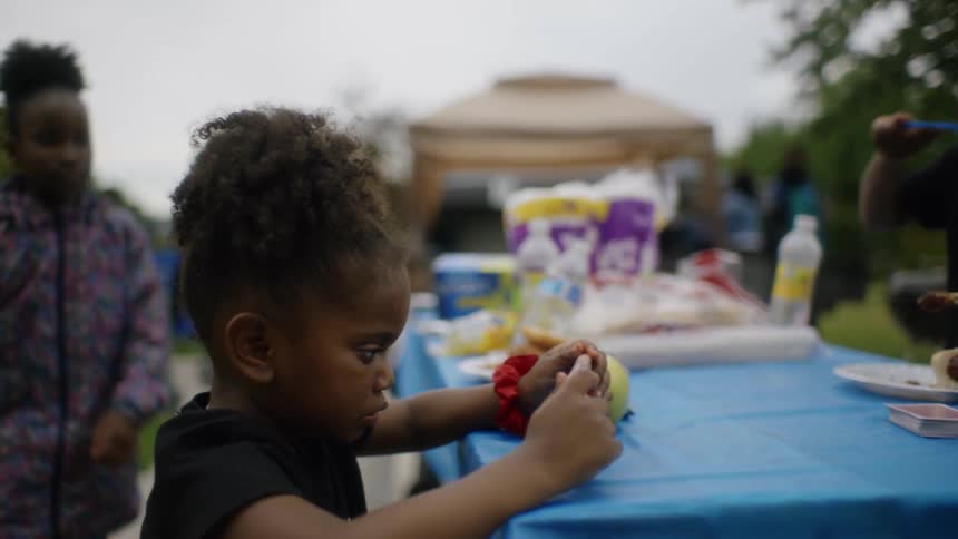 A little girl sits at a picnic table in a park and plays on a cell phone while her sister eats and another sister comes running and sits down at the table