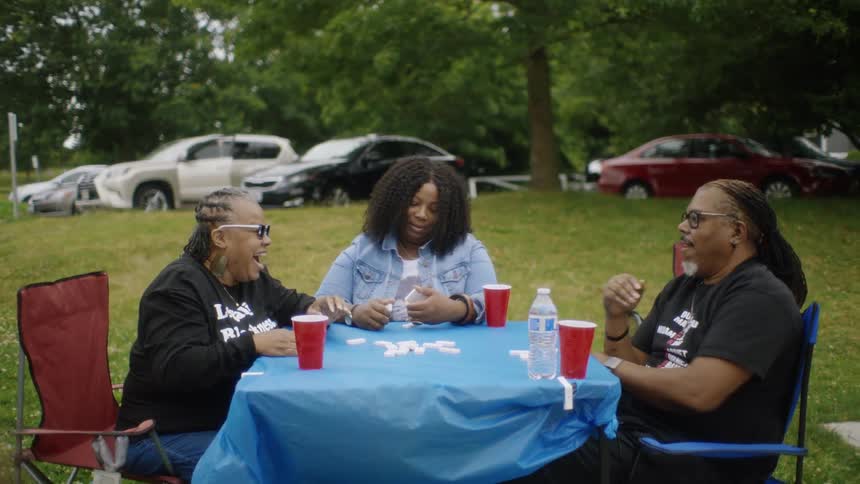Man with glasses and black shirt plays okey with women with dreadlocks curly hair denim jacket and black sweater sitting at table in public park
