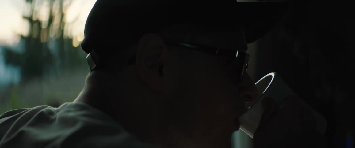 Man with black cap and glasses sits at table looking out the window and drinking from cup