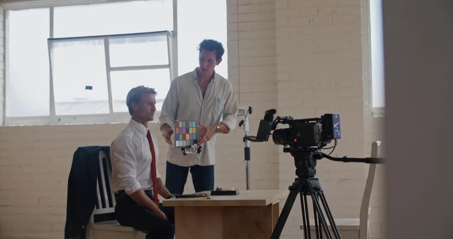 A man sits at a table while another gets camera equipment ready