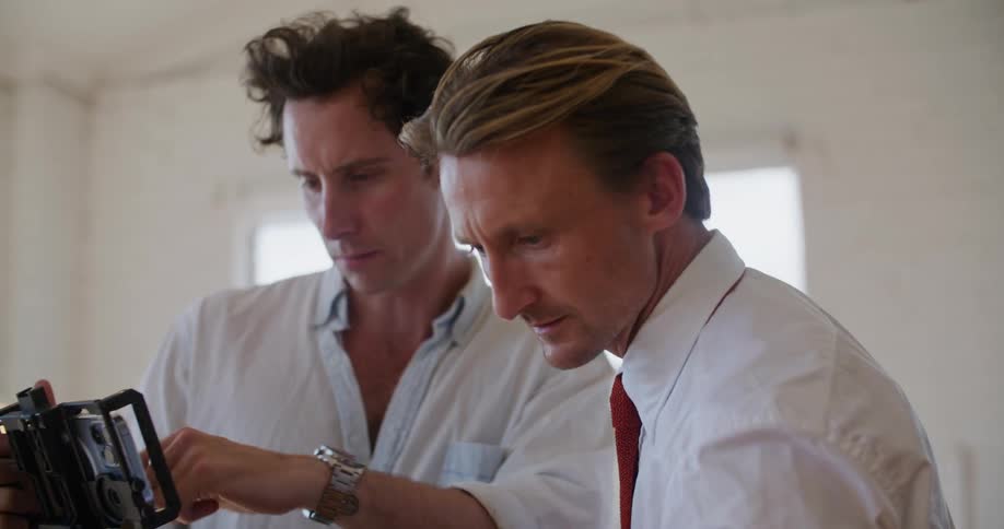 Two filmmakers standing in front of a camera talk while setting up a shot inside a white room