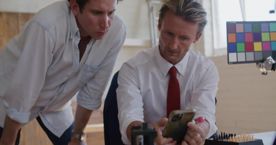 A man sitting at a desk in front of a video camera shows another information on his smartphone