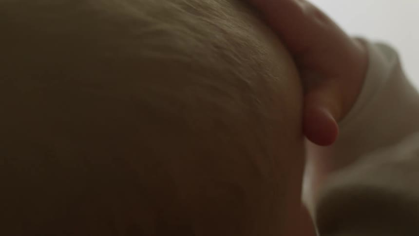 A baby with a pacifier in its mouth touches its own head with its hand
