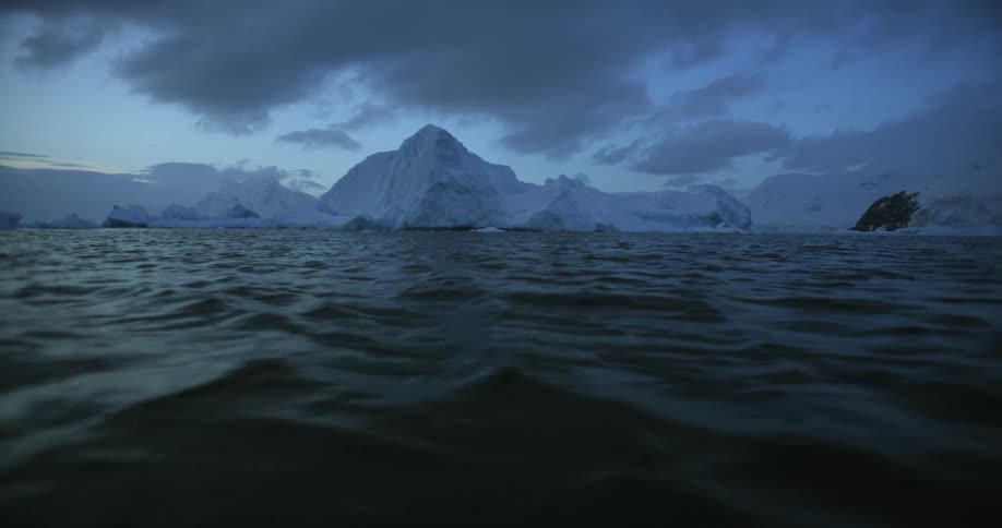 An iceberg stands tall in rough water under a cloudy sky