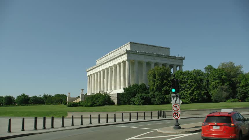 Pedestrians move along sidewalk and cars move along road near government memorial building