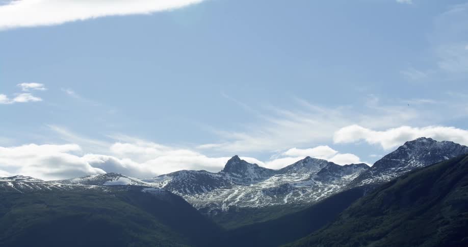 A mountain range covered in light snow and a bird flies across the cloudy sky