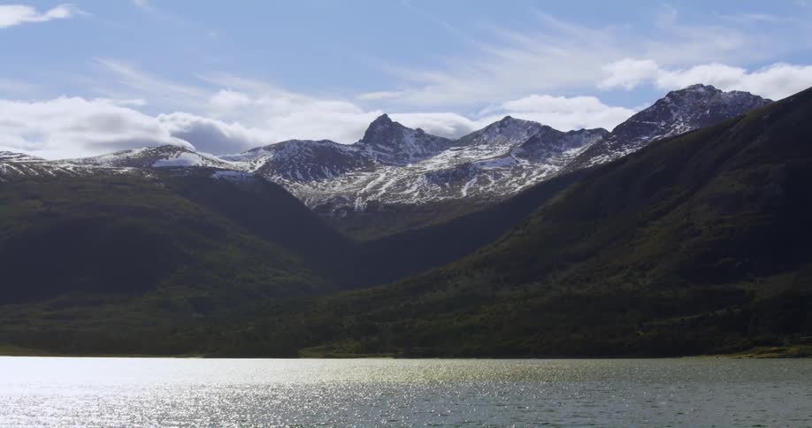 The sun reflects on the water as it ripples and waves and there are snow covered mountains in the distance under a cloudy sky