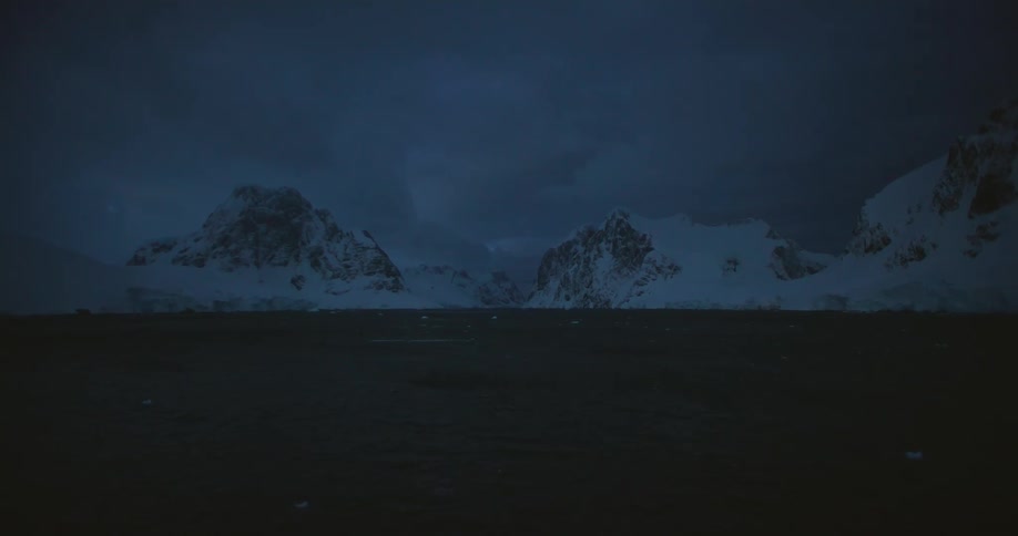 A dark body of water with snow covered rocky mountains under a cloudy gray sky