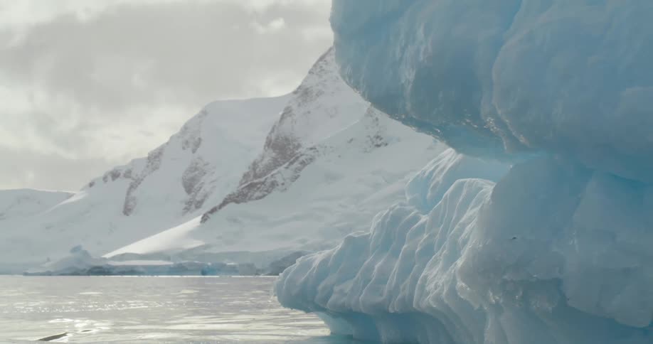 A melting iceberg is floating in the water and snow covered mountains are in the distance