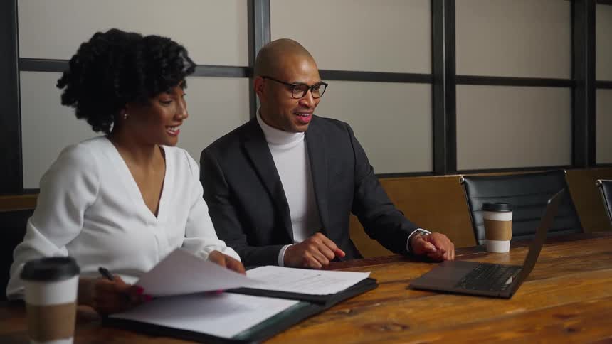 A man and woman sit in an office and wave at someone on a laptop