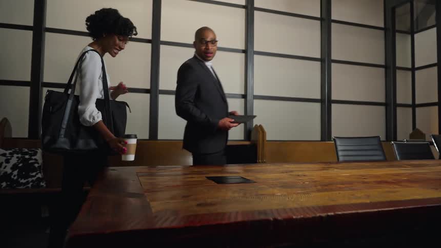 A man and woman walk in to an office and the woman sets a coffee cup down on the desk and a man sets his laptop down