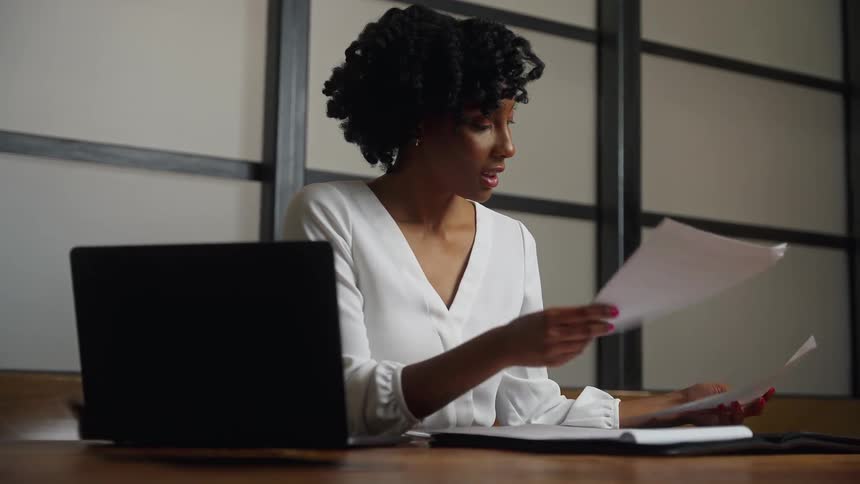 Woman sits at desk going through paperwork and using a laptop