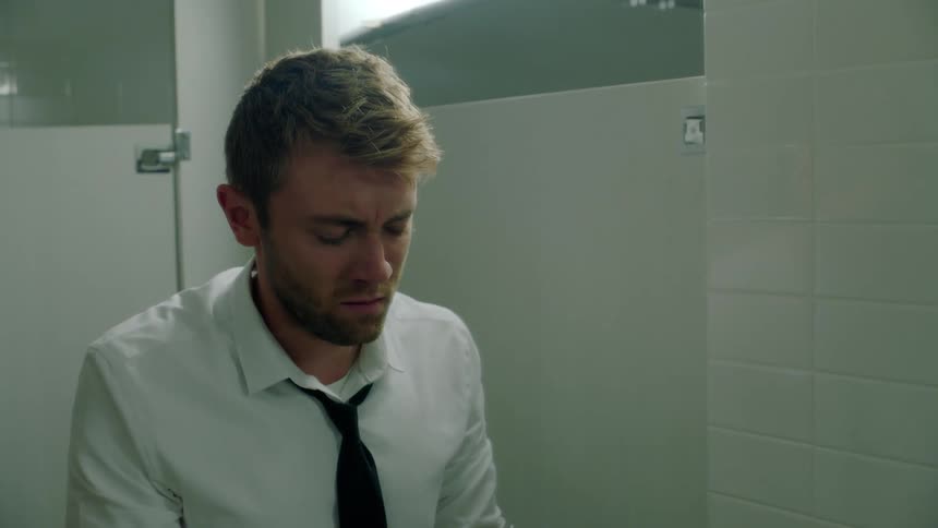 A man standing in a public bathroom looking distraught staring at the open beer bottles on the counter