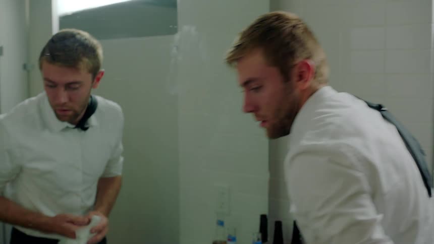 A man throws his tie over his shoulder before washing his hands and face in a public restroom
