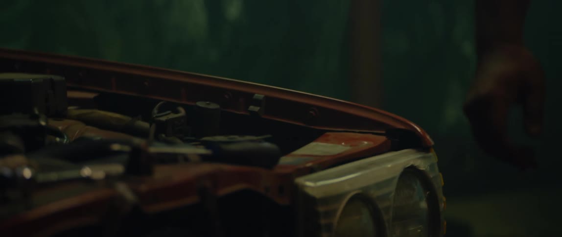 A man walks around to the front of his car at night and bends over the open hood