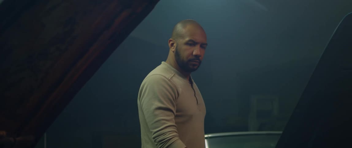 Bald bearded man walks around a car parked inside a dimly lit garage