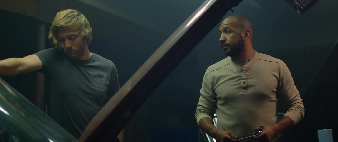 Two mechanics talk while working on the engines of a car parked inside a dimly lit garage