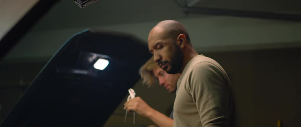 A man is working under the hood of a car and he hands pliers to a man working on a car next to him