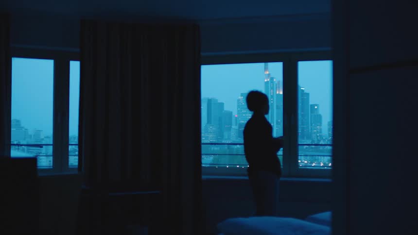 A woman walks across a room carrying a cup of coffee and looks out at the city skyline