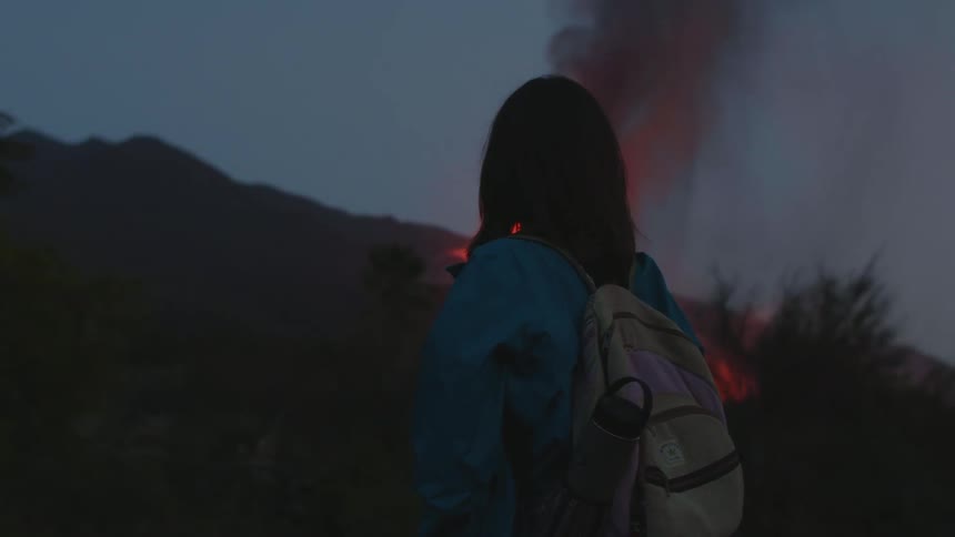 Woman with blue raincoat and gray backpack watches lava erupting at dusk
