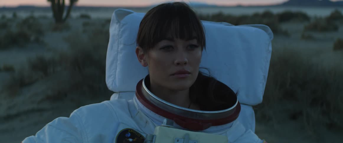 A woman wearing an astronaut suit and holding her helmet is standing in the desert