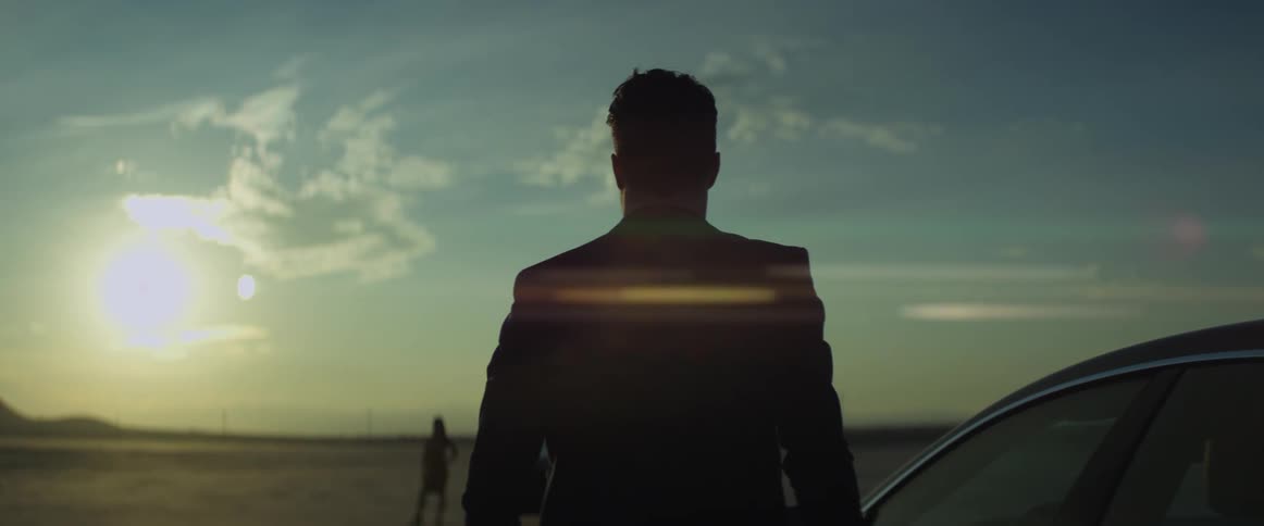 A woman is standing in the desert next to an automobile looking at the sun shining brightly over the mountains and a man gets out of a car parked nearby and walks toward her