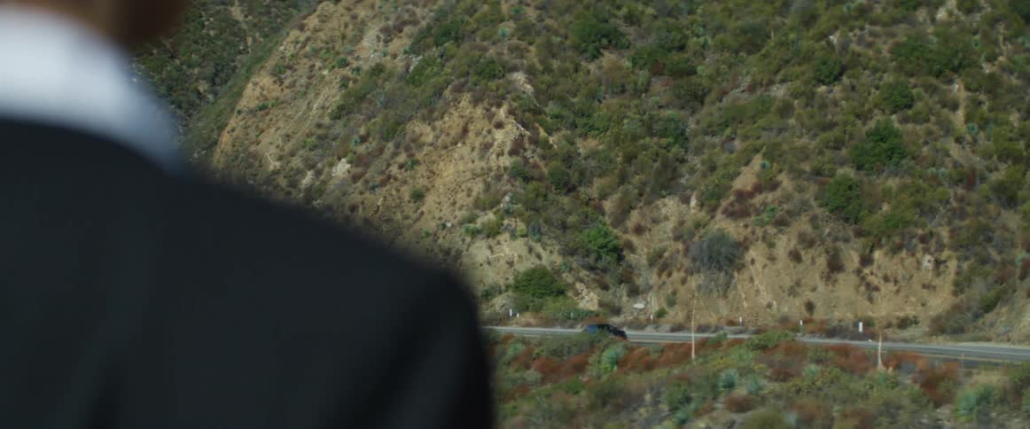 A person watches a grey car travel on the road next to the mountain