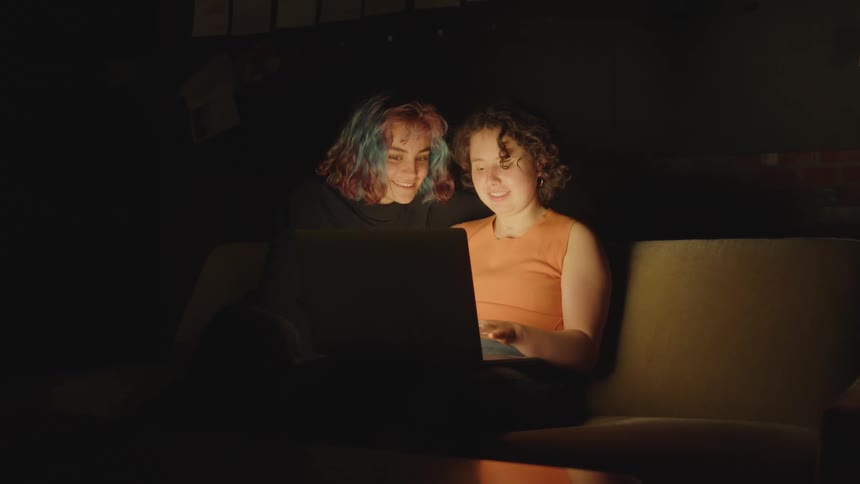 A woman smiles while sitting and talking to her daughter who is typing on a laptop