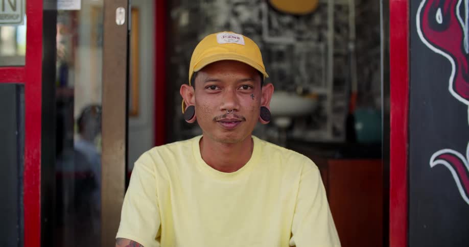 Man wearing a yellow hat and with earrings and a nose ring smiles while in front of a glass door