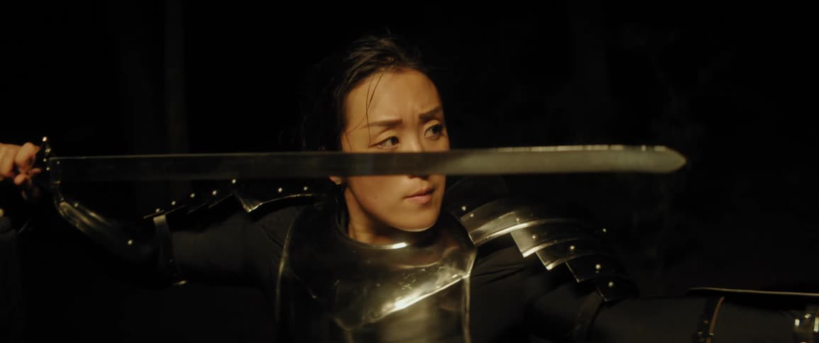 An asian woman is wearing armor and holding a sword near her face and her other arm out in front of her waiting to defend herself