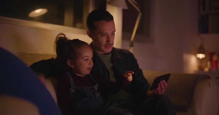 Man sits on a sofa with his daughter and they watch something on his mobile phone