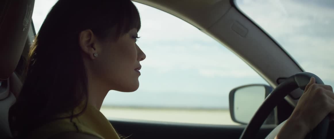 A woman briefly looks in her rear view mirror as she drives her automobile across a desert and there are mountains in the distance