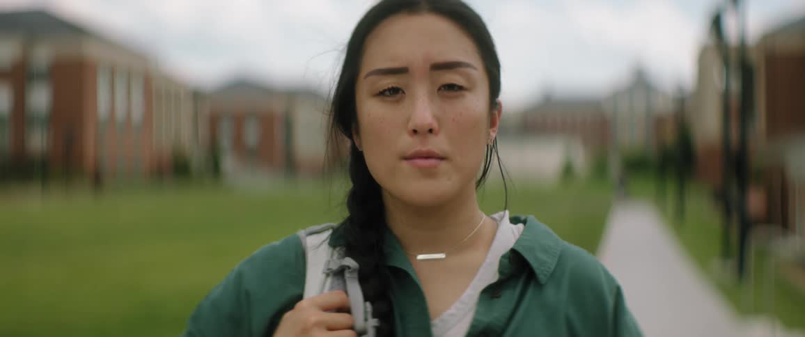 A college girl is standing outside of buildings on a sidewalk holding the strap of her backpack over her shoulder