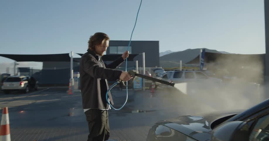 A man uses a sprayer to wash his car in a car wash bay on a sunny day