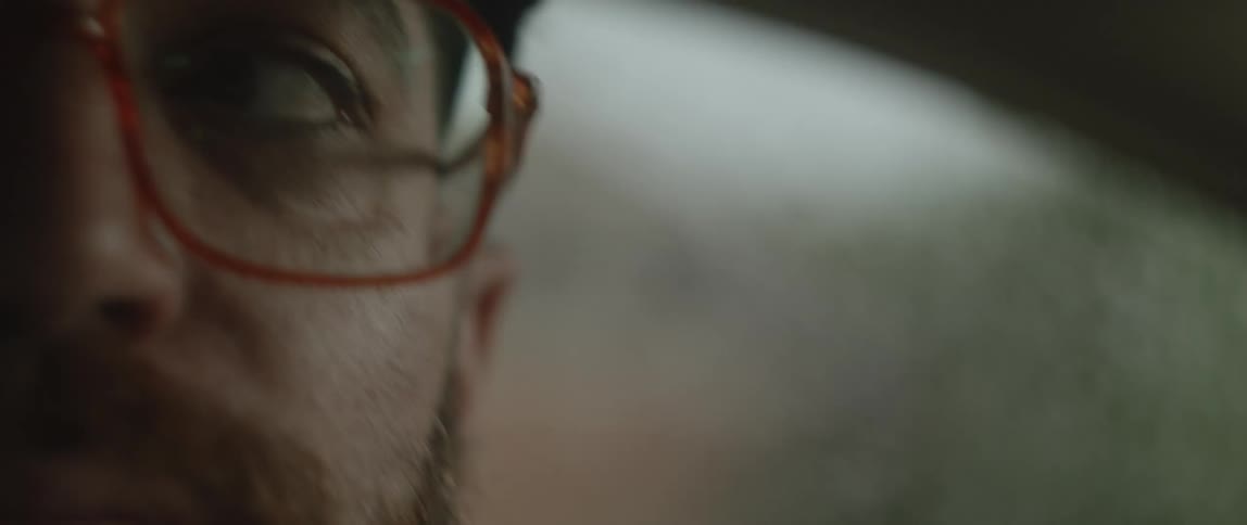 A bearded man wearing glasses looks around while driving in his car on a rainy day