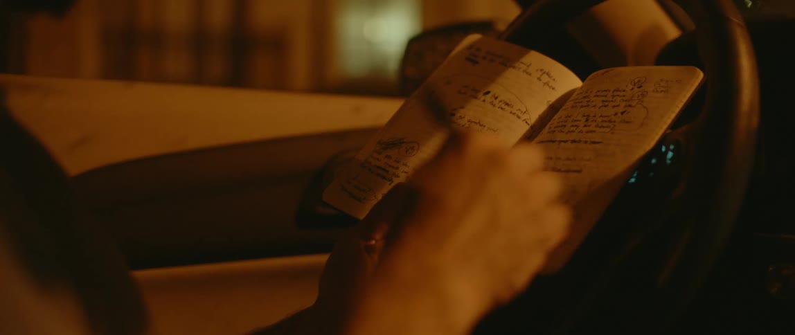 A man sits in a car and flips through a book of hand written notes while holding a pen