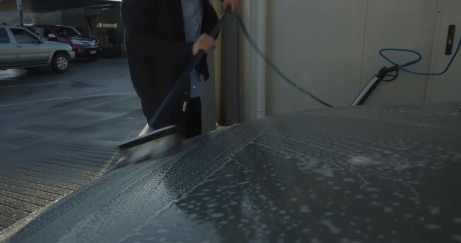 A man is outside at a car wash holding a soapy brush and washing his car