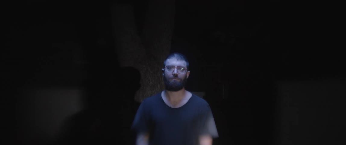 Man with a beard and wearing glasses stands in front of a tree as lights flash on him at night