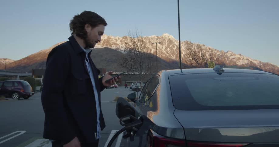 A man removes the fuel cap from his vehicle and inserts a gas pump before looking at his phone