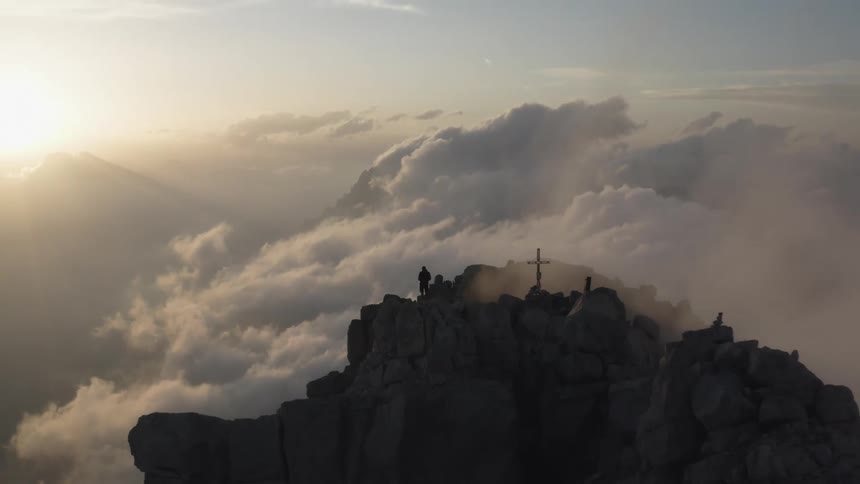 A man is standing on a mountain peak near a cross as the sun shines brightly over the clouds