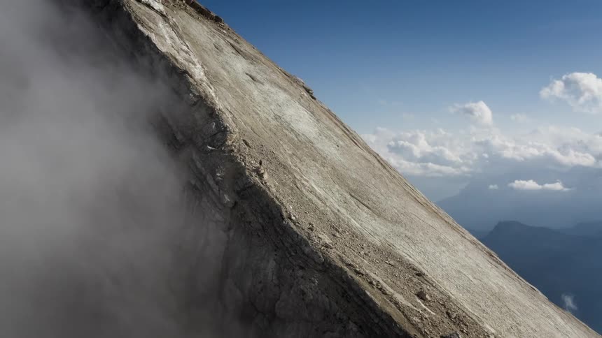 Fog is rising from the side of a mountain near where a person is standing and there are mountains in the distance high among the clouds
