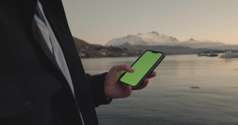 A man is standing near a lake holding a cell phone with a green screen and there are boats in the water and mountains in the distance