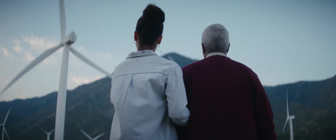 A woman points up at a spinning wind turbine while guiding an elderly man on a walk through a wind farm