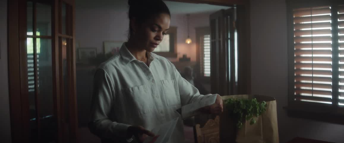 A young woman is carrying a bag of groceries through a room and she sets it on a table and then looks though the mail and opens a letter