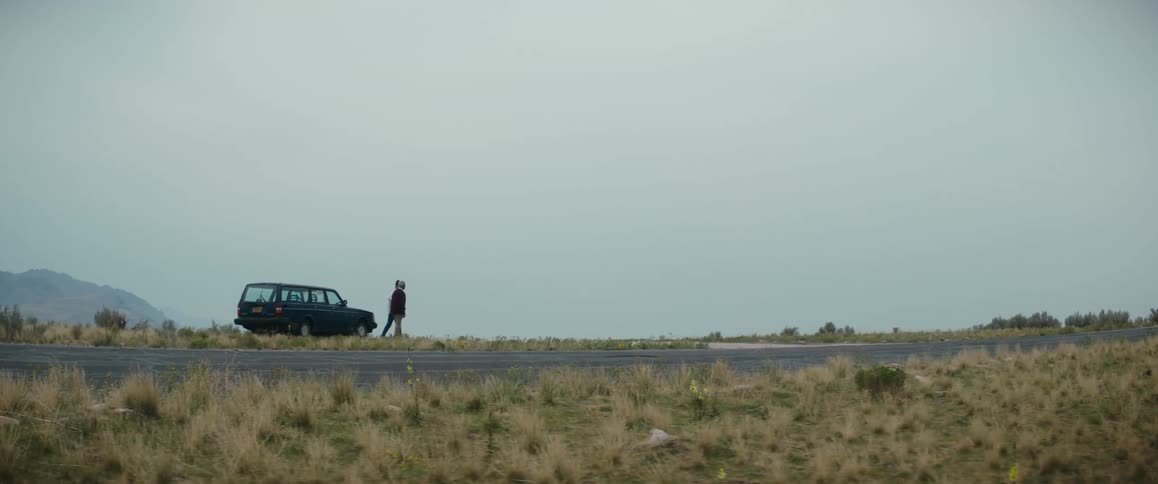Woman with bun and white shirt stands next to man with black blazer shirt and pants near car in the field