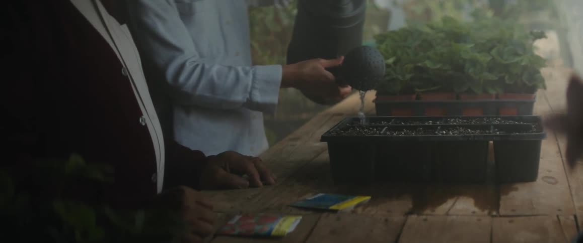 An old man stands next to a woman as she waters seeds in a planter