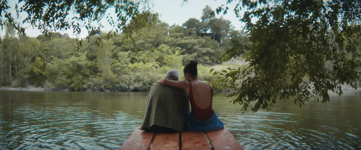 An elderly man and a young woman are sitting on a picnic table at the edge of a lake with woods on the other side as she puts her arm around him