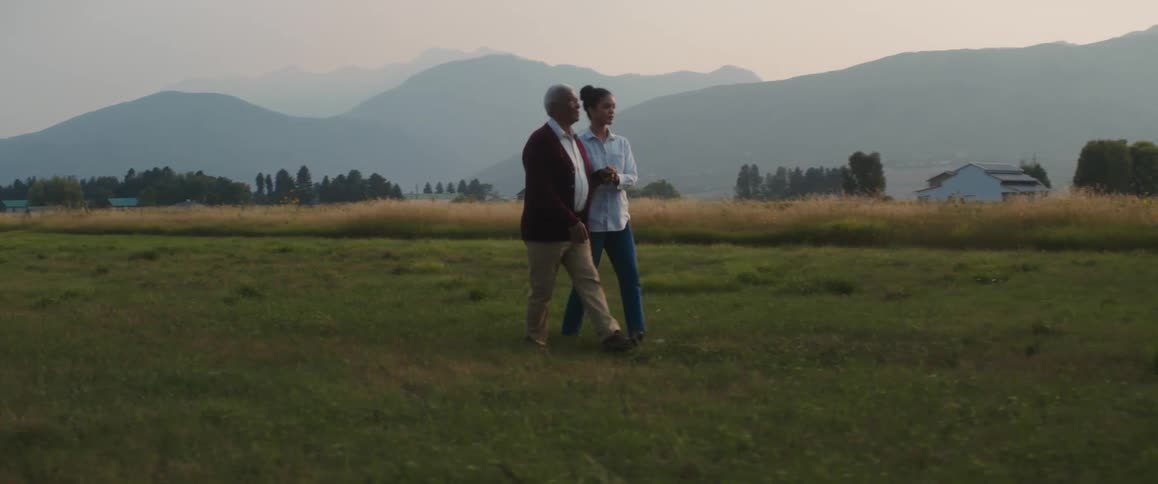 A woman guides her blind grandfather from their parked car across the farm land grass