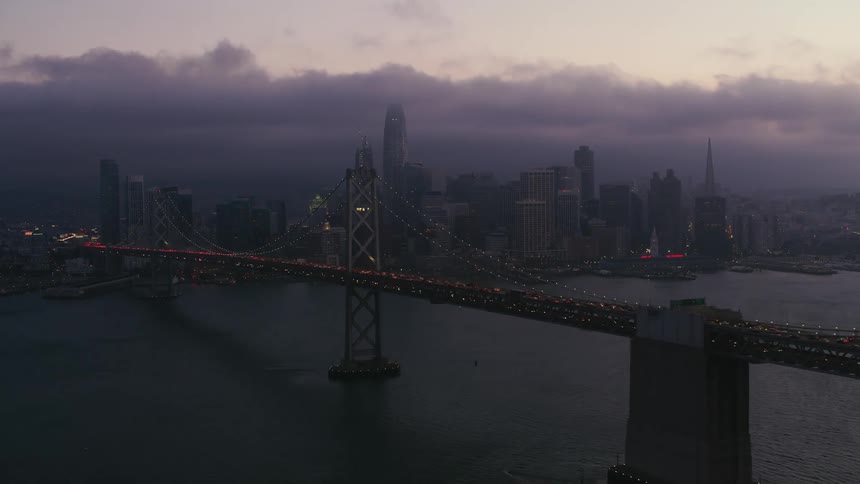 Traffic is moving across a suspension bridge as boats sail in the water below and towers and skyscrapers in the big city are standing tall in front of dark clouds
